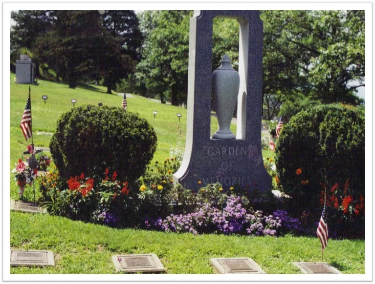 The Garden of Memories monument at Wildwood Cemetery, a tall granite arch holding a carved urn, flanked by clipped bushes and a bed of marigolds and petunias, with bronze markers set flush in the grass in front